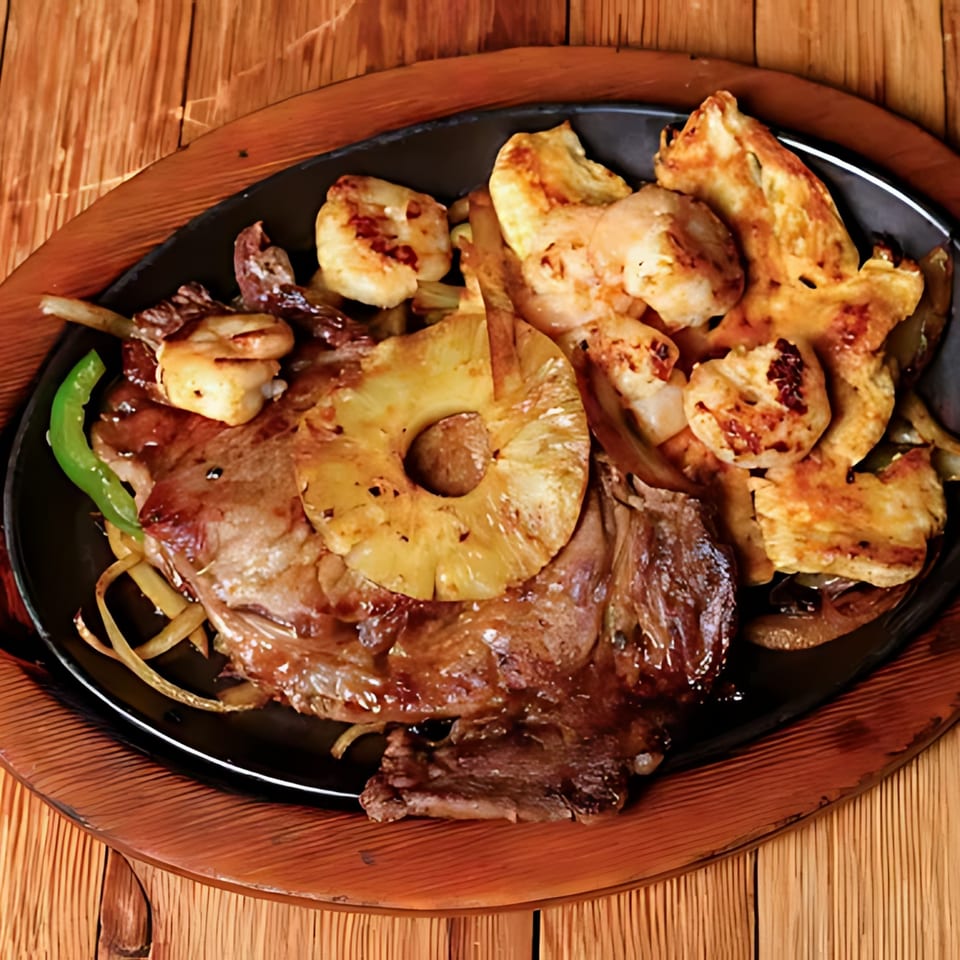 Sizzling Tequila Mexican Restaurant fajitas with steak, shrimp, pineapple, onions, and green peppers on a black platter.